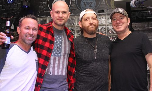 Four men smiling together at a club, one holding a beer.