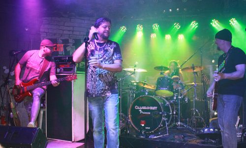 Band plays live onstage with singer at center under colorful lights.