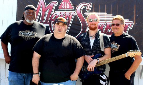 Four men pose in front of a Main Street banner, one holding a guitar.